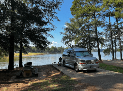 A silver van parked by a lake, surrounded by trees, with a person sitting nearby enjoying the view.