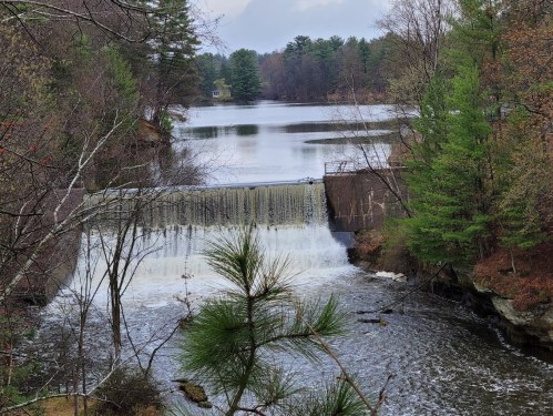 A serene view of a waterfall cascading over a dam into a calm lake, surrounded by trees and a cloudy sky.