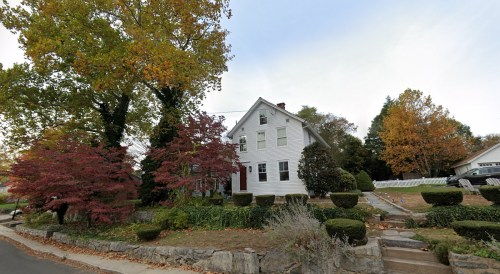 A white two-story house surrounded by colorful trees and neatly trimmed bushes on a quiet street.