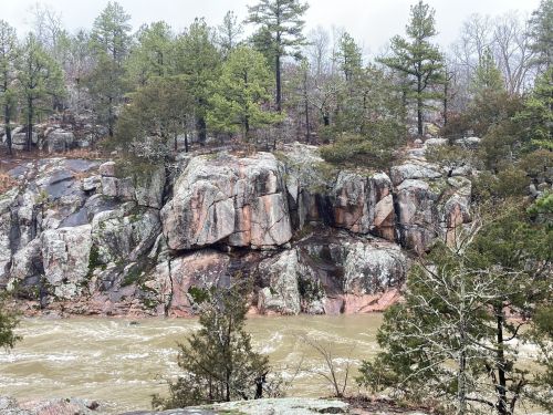 Rocky riverbank with large boulders and trees, surrounded by a misty, overcast sky. Water flows gently nearby.