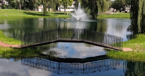 A serene pond with a fountain, featuring a black railing bridge reflecting in the water surrounded by greenery.