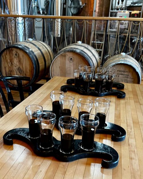 A wooden table with flight trays of dark drinks, barrels in the background, and brewing equipment visible.