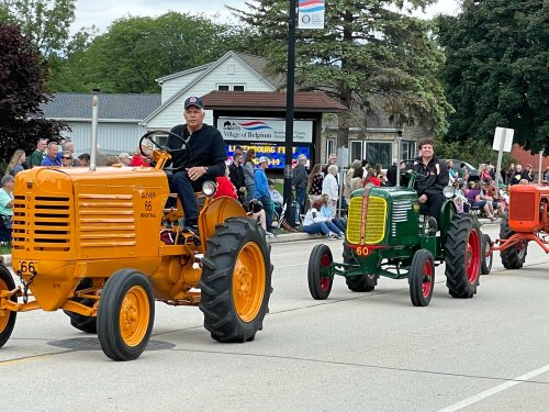 Two vintage tractors in a parade, one orange and one green, with riders smiling as they pass a crowd and buildings.
