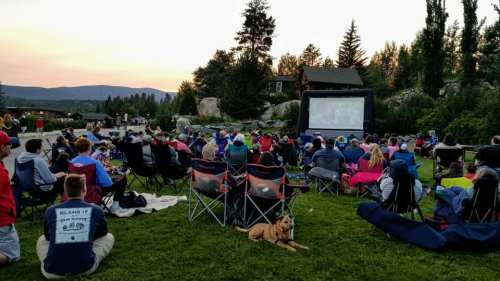 A crowd sits on the grass watching a movie on a large outdoor screen at sunset, with trees and mountains in the background.