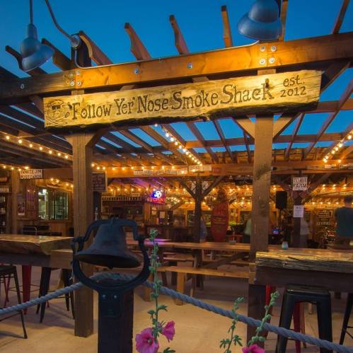 A rustic outdoor smoke shack with string lights and a wooden sign reading "Follow Yer Nose Smoke Shack est. 2012."