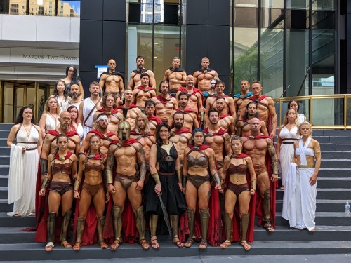 A large group of people in ancient Greek-inspired costumes, posing together outside a modern building.