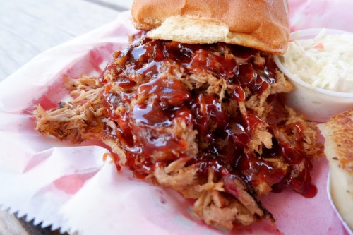 A close-up of a pulled pork sandwich topped with barbecue sauce, served with a side of coleslaw.