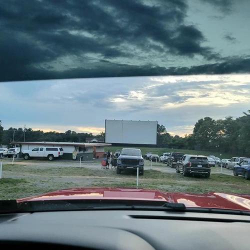 View from a car dashboard at a drive-in theater, with parked cars and a blank movie screen against a cloudy sky.