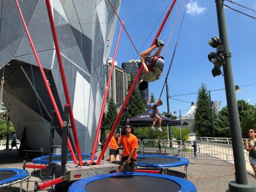 A person flips upside down on a trampoline while others watch and assist in a sunny outdoor setting.