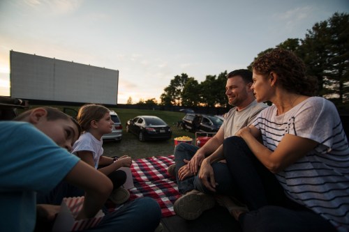 A family enjoys a picnic at a drive-in movie, sitting on a blanket with a screen in the background and cars parked nearby.