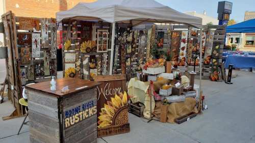 A craft booth featuring wooden decor, fall-themed items, and a "Welcome" sign under a canopy at an outdoor market.