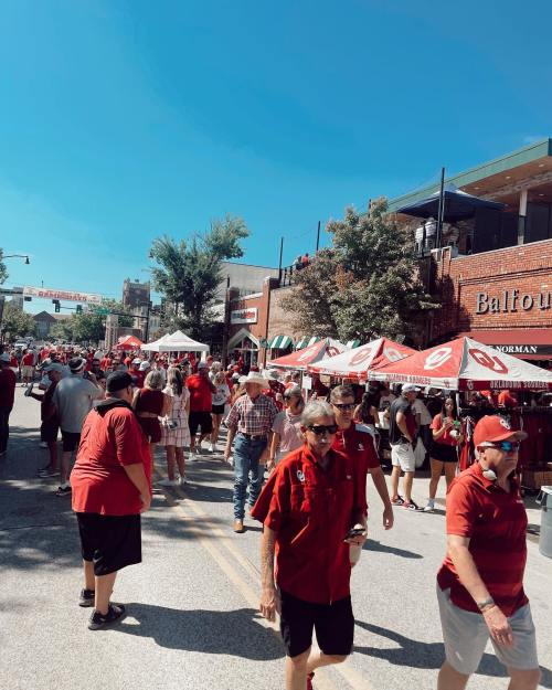 Crowd of people in red shirts walking down a street lined with tents and buildings on a sunny day.