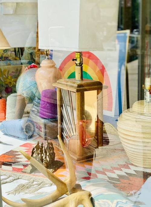 A colorful display of vintage decor, including a lamp, yarn, and art, seen through a shop window.