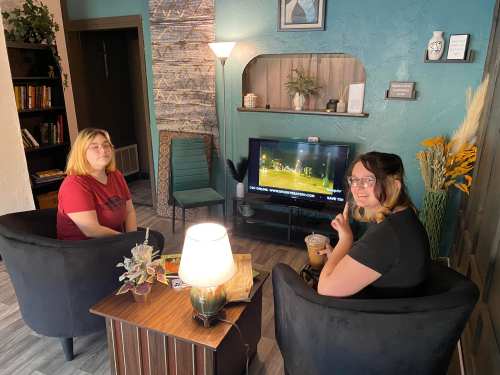 Two women sit in a cozy living room, one holding a drink, watching TV, with plants and bookshelves in the background.