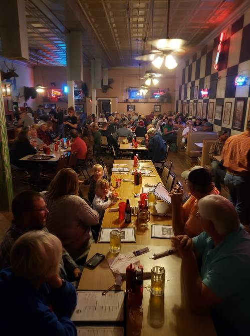 A busy restaurant filled with diners at long tables, colorful neon lights, and a cozy atmosphere.
