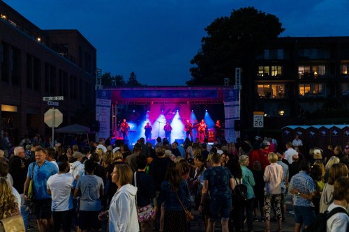 A lively outdoor concert scene with a crowd enjoying a band on stage, illuminated by colorful lights at dusk.