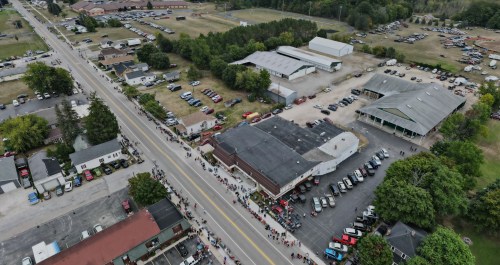 Aerial view of a busy street with a crowd, parked cars, and buildings in a rural area.