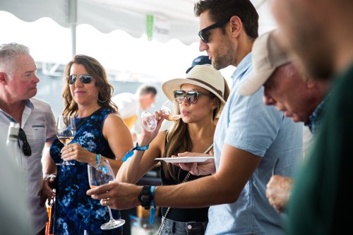 A group of people enjoying drinks at an outdoor event, with one woman sipping wine and another holding a plate.