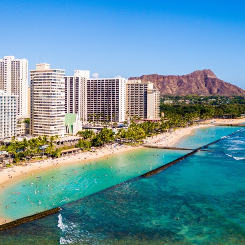 Aerial view of a beach with turquoise water, palm trees, and high-rise buildings against a mountain backdrop.