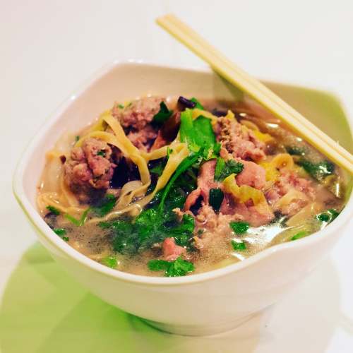 A bowl of soup with noodles, greens, and meat, garnished with herbs, served with chopsticks.