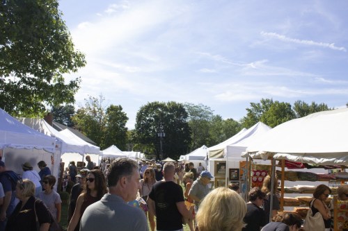 A bustling outdoor market with white tents, people browsing, and a clear blue sky overhead.