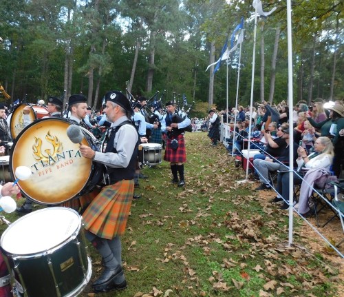A pipe band performs outdoors, with drummers in kilts and a crowd enjoying the event among fallen leaves.