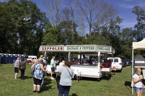A food stand offering zeppoles, sausage and peppers, and funnel cakes, with people lined up in a grassy area.
