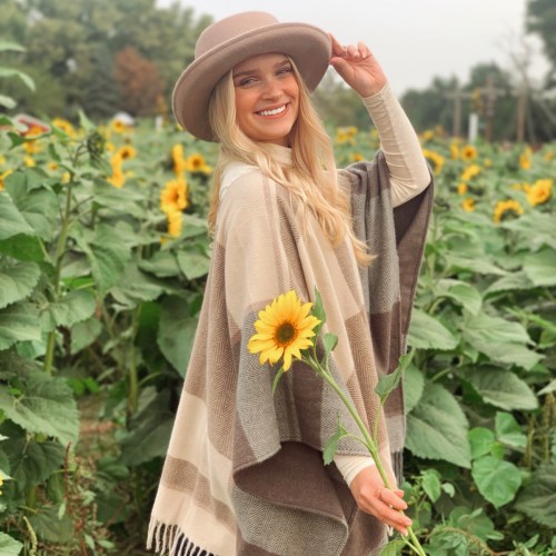 A smiling woman in a hat stands among sunflowers, wearing a cozy poncho and holding a sunflower.