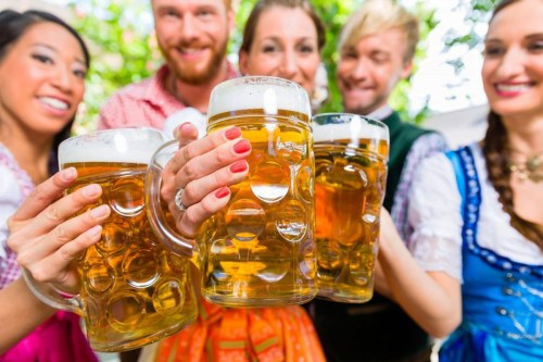 A group of five people in traditional attire cheerfully holding large beer mugs outdoors.