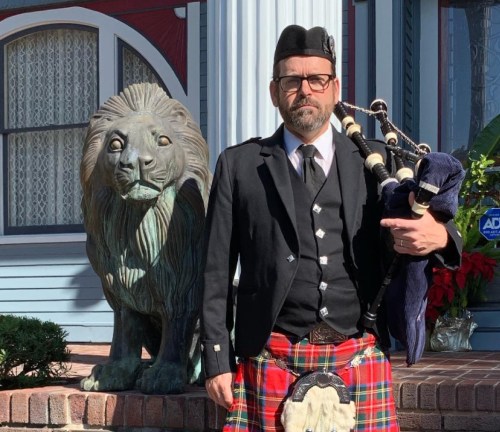 A man in a kilt and jacket stands next to a lion statue, holding a bagpipe, in front of a decorated house.