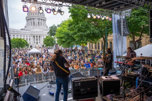 A lively outdoor concert scene with a large crowd in front of a stage, featuring musicians and a historic building in the background.