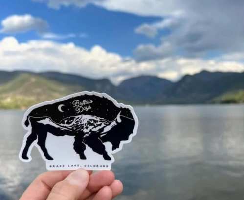 A hand holds a sticker of a buffalo silhouette against a scenic lake and mountain backdrop in Grand Lake, Colorado.