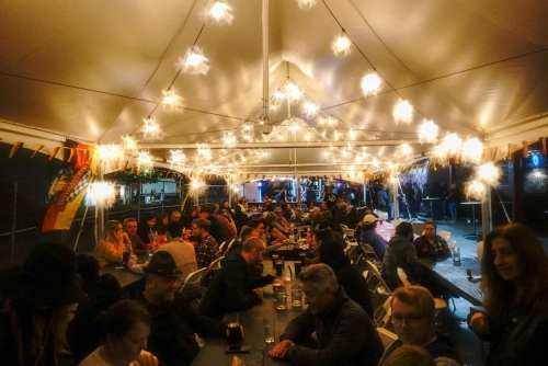 A lively outdoor gathering under a tent, filled with people enjoying drinks and string lights illuminating the scene.