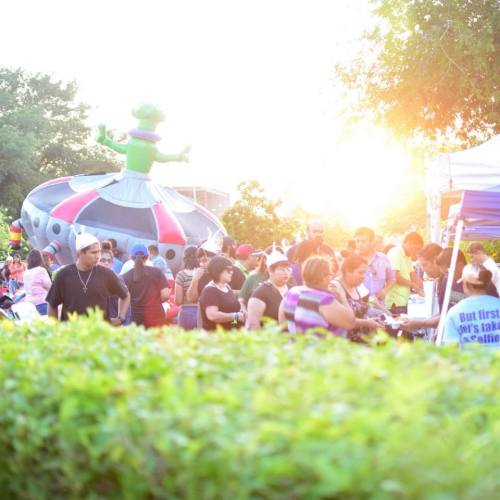 A lively outdoor event with people gathered around a colorful inflatable alien and food stalls, bathed in warm sunlight.