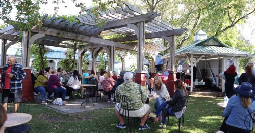 A lively outdoor gathering under a pergola, with people seated at tables enjoying the event in a green park setting.