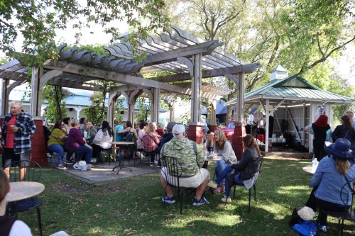 A lively outdoor gathering with people seated at tables under a wooden pergola, enjoying a sunny day.