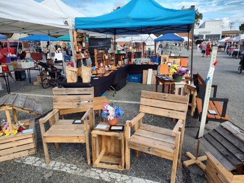 Wooden chairs and tables displayed at an outdoor market under blue tents, with various crafts and decorations in the background.