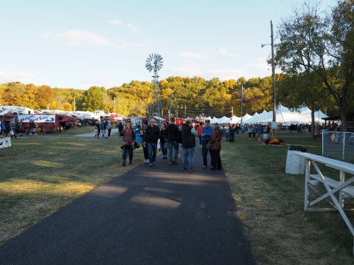 A crowd walks along a path at an outdoor fair, with tents and autumn trees in the background.