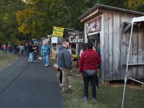A bustling outdoor market scene with people chatting near wooden stalls selling coffee and lemonade. Autumn foliage in the background.