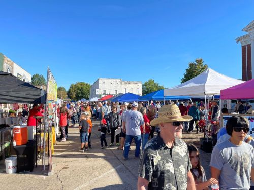 A bustling outdoor market scene with crowds, colorful tents, and clear blue skies. People are enjoying the event.