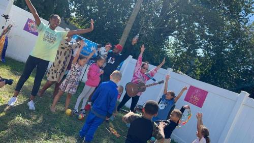 A group of children and adults joyfully participating in an outdoor event, with music and activities in a sunny setting.