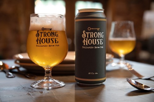 A glass and can of Strong House beer on a wooden table, with plates and utensils in the background.