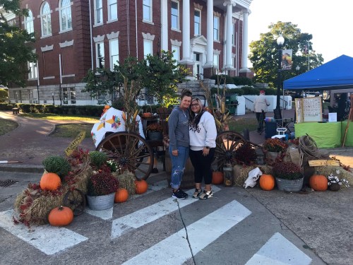 A couple stands in front of a decorated fall display with pumpkins and flowers, near a historic building.