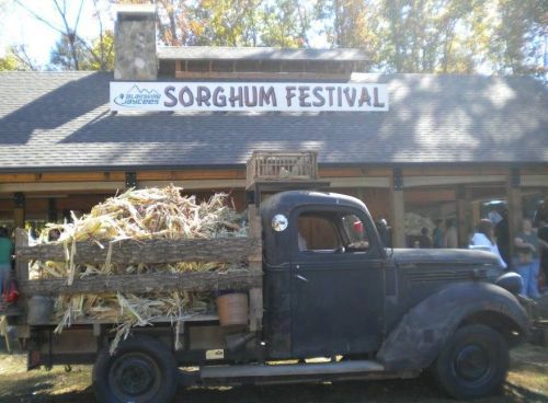 A vintage truck loaded with hay parked in front of a building with a "Sorghum Festival" sign.