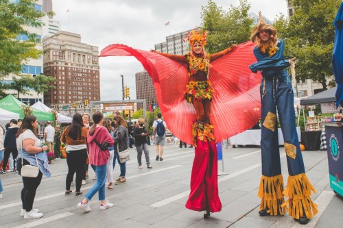 Two performers on stilts, one in a vibrant red costume and the other in a colorful outfit, entertain a crowd at an outdoor event.