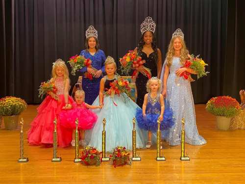 A group of young girls in colorful gowns and tiaras, holding flowers and trophies, posing together on stage.