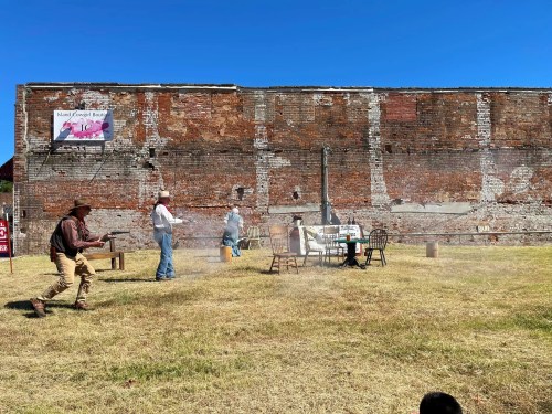 A western-themed scene with people in cowboy attire engaging in a mock shootout near a weathered brick wall.