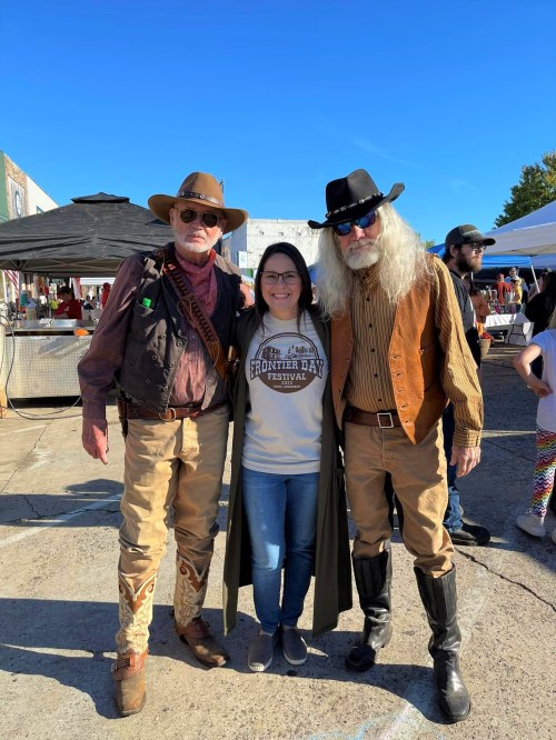 Two men in cowboy attire pose with a woman in a t-shirt at an outdoor festival under a clear blue sky.