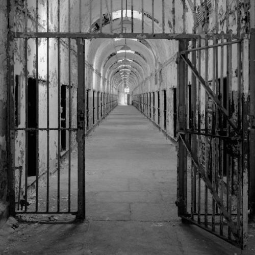 A dimly lit, abandoned prison corridor with barred gates and peeling walls, leading to a distant archway.