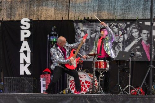 A lively band performs on stage, featuring a guitarist in a silver jacket and a drummer in a pink hat.